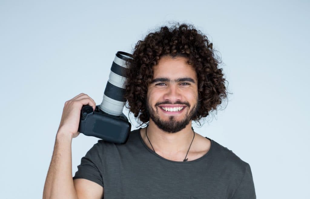 A smiling man resting a camera with a telephoto lens on his shoulder