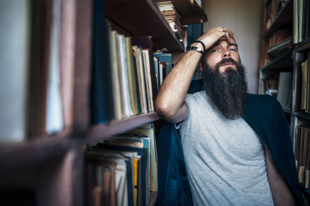 A bearded man leaning against a wall of library bookshelves