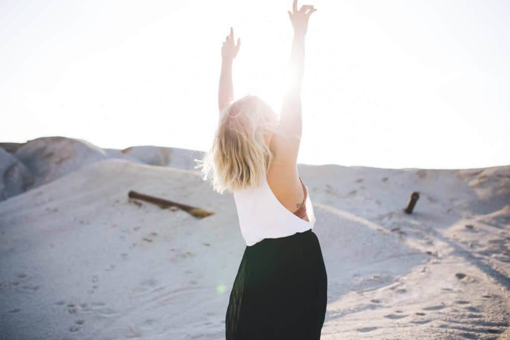 A person standing in pale sand dunes with both arms raised to the sun
