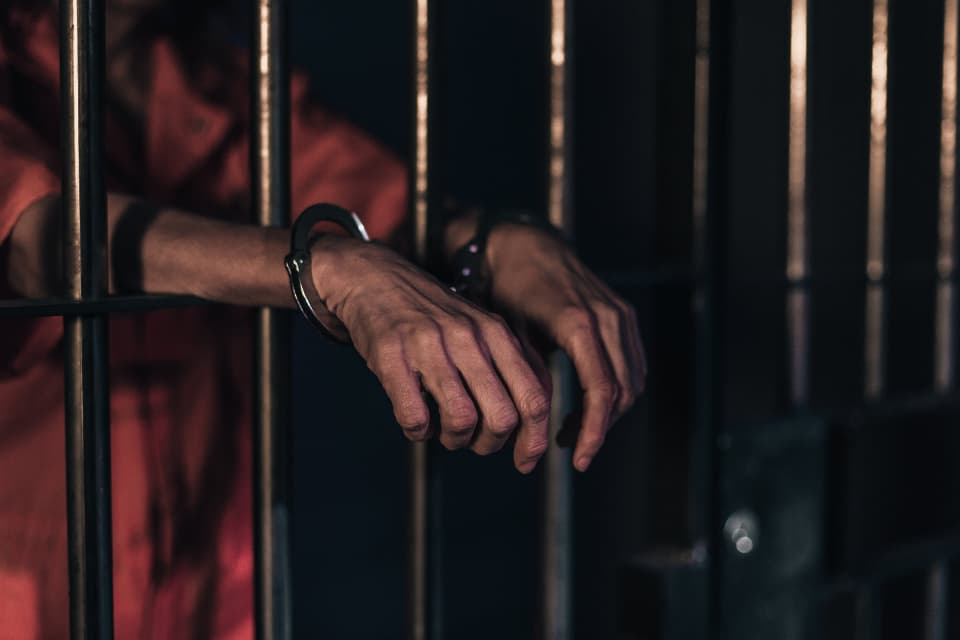 A pair of handcuffed hands resting on the bars of a jail cell