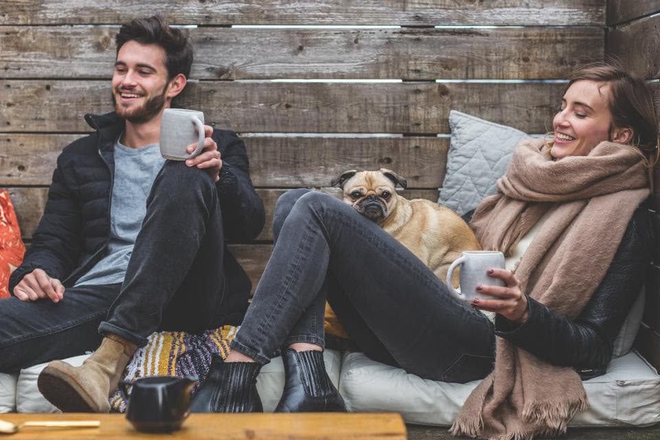 Two people sitting outdoors with mugs of coffee and a pug dog between them